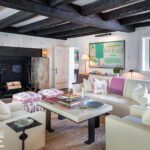 Cozy living room with exposed dark wood beams, a rustic stone fireplace, white paneled walls, comfortable beige sofas, colorful decor accents, and natural light streaming through a large window.