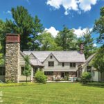 Rear exterior of a traditional Cotswold-style cottage featuring stone chimneys, clapboard siding, a spacious backyard with lush greenery, and outdoor seating under a bright blue sky with fluffy clouds.