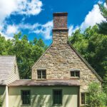 Side view of a Cotswold-style home showcasing a stone chimney, rustic facade, manicured landscaping, and lush greenery under a bright blue sky with scattered clouds.