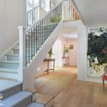 Elegant entryway featuring a staircase with a decorative wrought iron railing, light wood flooring, a large abstract painting, and natural light streaming through multiple windows.