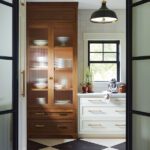Elegant butler's pantry featuring a mix of warm wood cabinetry with fluted glass doors and light sage green drawers with brass hardware. The space is complemented by a black-and-white checkered marble floor, a black-framed window letting in natural light, and a vintage-style pendant light, blending classic and modern design elements.