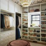 Luxurious walk-in closet featuring built-in white cabinetry, open shelving for shoes and accessories, and a central tufted velvet ottoman in a warm brown hue. The space is enhanced by a floral wallpapered ceiling, an elegant tiered chandelier, and a window providing natural light, creating a stylish and organized dressing area.