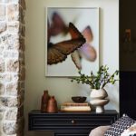 Stylish console vignette featuring a black geometric console table with gold hardware, adorned with a neutral vase holding green branches, ceramic decor pieces, and stacked books, set against a stone accent wall and framed artwork of a butterfly with soft lighting.