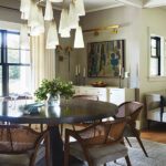Modern dining room featuring a round dark wood table surrounded by woven cane-back chairs with upholstered cushions, illuminated by an elegant multi-tiered chandelier with white shades. The space includes a sideboard with brass accents, abstract wall art, tall candlesticks, and a large window framed by neutral curtains, creating a sophisticated and airy atmosphere."