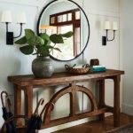 Entryway featuring a rustic wooden console table with decorative curved details, a large round mirror, sconces with cream shades, a ceramic vase with green foliage, and a woven basket for accents, complemented by a herringbone wood floor and a patterned rug.