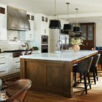 Spacious modern kitchen featuring a large wooden island with white quartz countertops, black pendant lighting, and upholstered barstools. The space includes white cabinetry with brass hardware, a custom range hood with brass detailing, glossy subway tile backsplash, and commercial-grade appliances. A cozy dining nook and built-in cabinetry complete the open and inviting design.