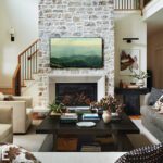 Bright and inviting living room featuring a stone fireplace with a mounted landscape TV, a black coffee table styled with books and vases, and a mix of neutral-toned seating, including a gray sofa and cream armchairs. The space is enhanced by layered textures, thoughtful decor, and a modern open layout with a staircase and a view of adjoining rooms.