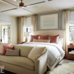 Serene primary bedroom featuring a plush upholstered bed with soft white linens, accentuated by blush and neutral throw pillows. The room includes matching wood nightstands with modern lamps, an elegant cream settee at the foot of the bed, and large windows dressed in light, flowing curtains.