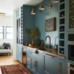 Charming scullery featuring custom teal cabinetry with brass hardware, a wood countertop, and integrated wine storage. The space is accented with a farmhouse-style sink, wall-mounted brass sconces, a colorful vintage rug, and modern artwork, creating a blend of functionality and timeless design.