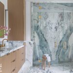 Elegant bathroom with book-matched marble walls and flooring, a glass-enclosed shower with gold fixtures, a double vanity with marble countertops, wooden cabinetry, and fresh floral accents.