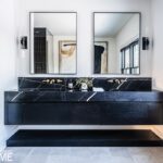 Modern bathroom with a black floating vanity featuring a dramatic black marble countertop and backsplash, gold fixtures, dual mirrors, minimalist sconces, and neutral tile flooring.