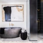 Luxury bathroom with a freestanding concrete bathtub, gold fixtures, abstract wall art, a black glass-enclosed shower with gold hardware, and neutral tile flooring.