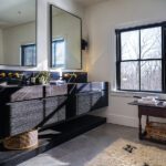 Stylish bathroom featuring a floating black vanity with a black marble countertop, dual gold faucets, large mirrors, a shag rug, a window with natural light, and modern accents.