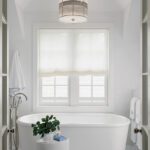 Elegant white bathroom with a freestanding soaking tub, geometric tile flooring, a large window with sheer roman shades, and a modern pendant light fixture.