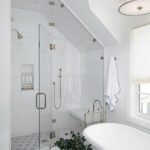 Modern white bathroom featuring a glass-enclosed walk-in shower with chrome fixtures, built-in shower niche, freestanding soaking tub, geometric tile flooring, and a statement pendant light.