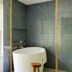Luxury bathroom featuring a sleek white freestanding bathtub against textured blue vertical tiles, accented with brass fixtures and a wooden stool. Modern spa-like retreat with minimalist design and calming ambiance.