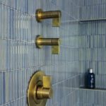 Modern shower design featuring brushed brass fixtures against elegant vertical blue ceramic tiles. Stylish bathroom interior with sleek controls and built-in shelf for toiletries. Luxury shower aesthetic.