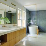 Modern luxury bathroom featuring a freestanding white bathtub, blue textured tile accent wall, double vanity with wooden cabinetry, gold-framed mirrors, and brass fixtures. Bright and airy design with natural light and greenery for a spa-like ambiance.