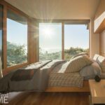 Sunlit modern bedroom with floor-to-ceiling windows offering breathtaking views of nature, featuring a cozy bed with layered bedding, light wood furniture, and minimalist decor.