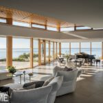 Spacious modern living area with floor-to-ceiling windows showcasing stunning ocean views, featuring plush seating, a grand piano, contemporary decor, and natural wood accents.