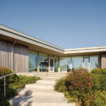 Modern home exterior with a minimalist design featuring large glass windows, a wooden facade, landscaped greenery, and a stone staircase leading to the entrance under a clear blue sky.