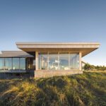 Contemporary coastal home with expansive glass walls and a flat roof design, set atop a grassy hillside with panoramic views of the surrounding landscape under a clear sky.