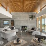 Stylish modern living room featuring a concrete accent wall with a built-in fireplace, cozy curved seating, a wood-paneled ceiling, and floor-to-ceiling windows showcasing scenic outdoor views.