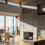Modern music room featuring a concrete fireplace, warm wood accents, floor-to-ceiling windows offering ocean views, and a cozy setup with guitars and music stands bathed in natural light.