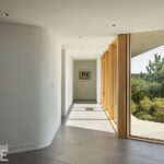 Bright modern hallway featuring curved walls, large floor-to-ceiling windows with views of lush greenery, and minimalist decor with a framed artwork at the end of the corridor.