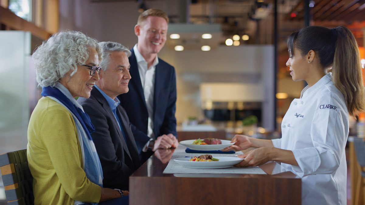 Chef at Clarke Showroom presenting gourmet dishes to a group of engaged visitors during a live kitchen demonstration.