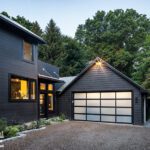 Side view of a contemporary home with a black exterior, a modern garage door, and a gravel driveway framed by trees and shrubs.