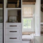 Charming built-in storage with white cabinetry, leather strap handles, exposed wooden beams, and a cozy window seat with natural light streaming through a Roman shade.