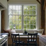 Bright and inviting dining nook with a window seat, farmhouse-style table, black chairs, and large windows offering lush garden views framed by rustic wooden beams.