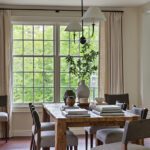 Elegant dining room with a rustic wooden table, gray upholstered chairs, neutral drapery, and large windows providing natural light and scenic outdoor views.