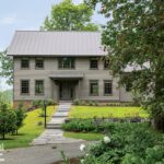 Picturesque home exterior featuring a classic design with a gray facade, metal roof, stone pathway, vibrant landscaping, and a backdrop of lush greenery.