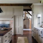 Rustic yet modern kitchen with wood-beamed ceilings, cream cabinetry, a large wooden island, farmhouse sink, and brick fireplace adding warmth and character.