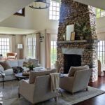 Spacious living room with a towering stone fireplace, neutral-toned furniture, large windows, and warm natural light creating a cozy and elegant atmosphere