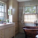Elegant guest bathroom with a copper soaking tub, natural stone tile walls, custom beige cabinetry, and windows showcasing serene outdoor views.