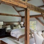 Cozy primary bedroom with exposed wooden beams, a neutral palette, a wood-frame bed, and a seating area, creating a warm and rustic yet sophisticated retreat.