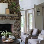 Living room featuring a towering stone fireplace, neutral-toned armchairs, and large windows with soft drapery, offering a blend of rustic and modern elegance.