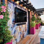 Outdoor entertainment setup on a rooftop deck featuring a large wall-mounted TV, colorful floral decor, and a wooden pergola.