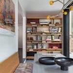 Modern dining area featuring a built-in wooden bookshelf filled with books and decor, colorful upholstered bench seating, a sleek gray dining table with black decorative bowls, and a contemporary chandelier with amber glass accents.