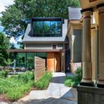Modern architectural home exterior showcasing a blend of classic and contemporary design elements, featuring large glass windows, a sleek wooden entrance, vertical wooden privacy panels, and lush landscaped greenery. The backdrop includes tall trees and a blue sky, enhancing the serene setting.