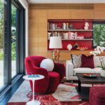 Modern family room with vibrant design elements, featuring a bold red accent chair, custom built-in wooden shelves with red backing, a white sofa with patterned throw pillows, a sleek black coffee table, and a large sliding glass door that provides natural light and views of the outdoors.