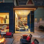 Stylish indoor-outdoor living space at twilight featuring a cozy fire pit surrounded by modern black chairs with red cushions, a glass-walled dining area with built-in bookshelves and warm lighting, and a view into an inviting kitchen with contemporary design. The stone patio and architectural details create a seamless blend of comfort and elegance.