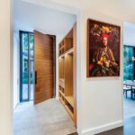 Elegant mudroom entryway featuring a modern wooden pivot door, built-in wooden storage cubbies, and a vibrant framed portrait on the wall. The space connects to a bright interior with large windows, showcasing a seamless blend of functionality and artistic decor.
