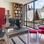 Bright and modern home office featuring a sleek desk with dual monitors, a stylish upholstered office chair, a vibrant red lounge chair with an ottoman, a floor-to-ceiling bookshelf, and large windows offering scenic views of a traditional rooftop and lush greenery.