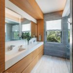 Modern primary bathroom featuring a double vanity with a sleek white countertop, warm wooden cabinetry, and large mirrors with integrated lighting. The design includes a spacious glass-enclosed shower, marble tile flooring, and a window with natural light and serene outdoor views.