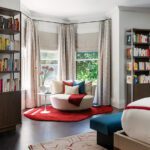 Cozy primary bedroom corner featuring a plush curved chair with colorful throw pillows, set on a vibrant red area rug. Surrounded by floor-to-ceiling windows with soft drapery, the space includes built-in bookshelves, dark wood flooring, and a contemporary bed with a patterned rug for a serene and inviting atmosphere.