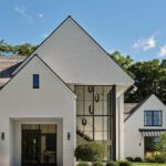 Modern agrarian-style front entrance of a white shingled Greenwich home with soaring glass windows and pitched gables under a clear blue sky.